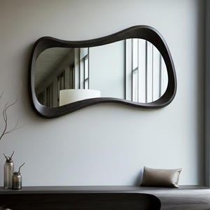 Könnte beinhalten: Ein moderner Spiegel mit dunklem Holzrahmen und einer ungewöhnlichen, fließenden Form hängt an einer hellgrauen Wand. Darunter eine passende dunkle Holzbank mit einer geschwungenen Aussparung und einem kleinen Kissen. Dekorative Glasflaschen und eine Vase mit Zweigen stehen auf der Bank.