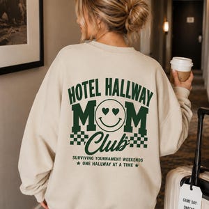 以下が含まれることがあります： ダークグリーンの「HOTEL HALLWAY MOM Club」の文字が入ったベージュのスウェットシャツ。ハートの目をしたスマイリーフェイスと市松模様のデザイン。背中には「SURVIVING TOURNAMENT WEEKENDS ONE HALLWAY AT A TIME」の文字もプリントされています。