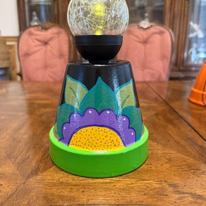 May include: A decorative lamp featuring a crackle glass globe on top of a painted flower pot. The pot is painted black with a green, purple, and yellow floral design, and a green base. The lamp is on a wooden table.