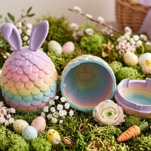 May include: A pastel-colored Easter egg-shaped container with bunny ears, open to reveal a rainbow interior. The egg is surrounded by moss, decorative eggs, flowers, and small chick and carrot figurines, creating a festive Easter scene.