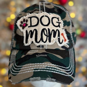 May include: Camouflage baseball cap with the words "DOG MOM" embroidered on a patch. The patch includes a paw print and a bone. The cap has a distressed look with white stitching.