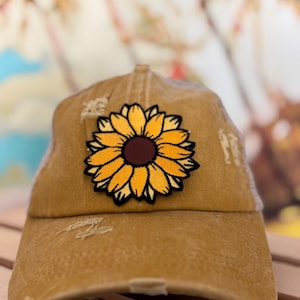 May include: Mustard-colored baseball cap with a distressed look, featuring a sunflower patch. The cap has a mesh back and a curved brim. The sunflower has yellow petals and a brown center. The cap is designed for casual wear.