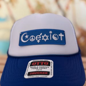 Może przedstawiać: Niebiesko-biała czapka typu trucker z napisem "Coexist" w niebieskim prostokącie. Słowo składa się z symboli różnych religii i filozofii. Czapka ma piankowy przód i zapięcie na zatrzask.