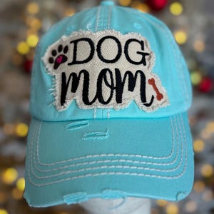 May include: A distressed light blue baseball cap with the words "DOG MOM" in black script and a paw print design. The cap has a frayed patch and a small bone graphic, with white stitching and a pre-curved bill.