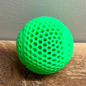 May include: A bright green ball with a honeycomb pattern. The ball is made of a solid material and sits on a wooden surface. The ball's design features numerous hexagonal openings throughout its surface.