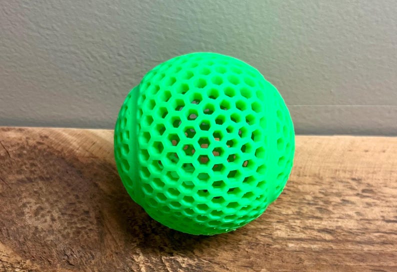 May include: A bright green ball with a honeycomb pattern. The ball is made of a solid material and sits on a wooden surface. The ball's design features numerous hexagonal openings throughout its surface.