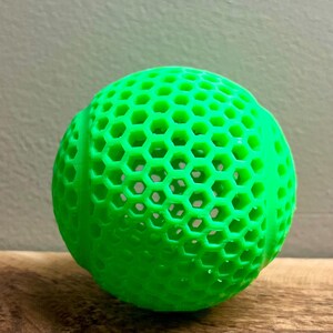 May include: A bright green, spherical object with a honeycomb pattern. The ball is made of a solid material and has a textured surface. The object is sitting on a wooden surface.