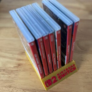 Op de afbeelding: Een gele Nintendo Switch game cartridge houder met rode letters. De houder is gevuld met verschillende game cases met rode ruggen. De houder staat op een houten oppervlak.