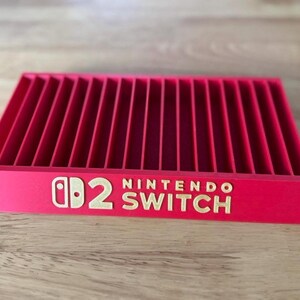 Op de afbeelding: Een helderrode Nintendo Switch game cartridge houder. De houder heeft meerdere verticale sleuven en is voorzien van het Nintendo Switch logo in gouden letters. Het ontwerp is rechthoekig en staat op een houten oppervlak.