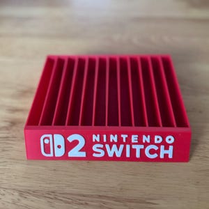 Puede incluir: Un soporte rojo para cartuchos de juegos de Nintendo Switch. El soporte es rectangular con ranuras verticales y el logotipo de Nintendo Switch en blanco. El soporte es de plástico y está sobre una superficie de madera.