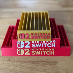 May include: A red and yellow Nintendo Switch game cartridge holder. The holder has the Nintendo Switch logo and the number 2 printed on it. The holder is made of plastic and has multiple slots for game cartridges.