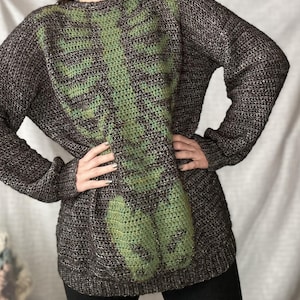 Puede incluir: Un suéter gris oscuro con un diseño de esqueleto de ganchillo verde. El suéter de manga larga tiene un patrón de punto texturizado y cuello redondo. El diseño del esqueleto está centrado en la parte delantera del suéter.