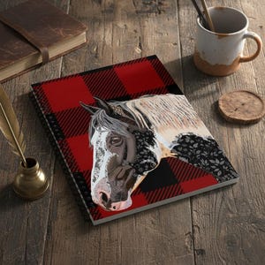 Puede incluir: Cuaderno de espiral con una ilustración de la cabeza de un caballo sobre una cubierta a cuadros rojos y negros. La cara del caballo es una mezcla de blanco, negro y marrón, con un estampado floral en la melena. El cuaderno está sobre una superficie de madera.