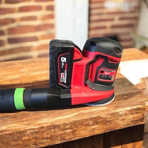 May include: A red and black cordless sander with a black battery pack. The sander has a round sanding pad and a black dust collection port with a green ring. The battery pack reads "5.0" and "M18 REDLITHIUM".