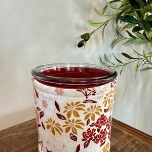 Op de afbeelding: Een heldere glazen pot met een bordeauxrode binnenkant, omwikkeld met een stoffen hoes. De hoes heeft een bloemenpatroon met witte, gouden en bordeauxrode bloemen op een crèmekleurige achtergrond. De pot staat op een houten oppervlak.