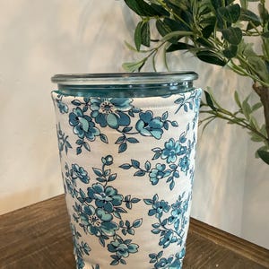 Op de afbeelding: Een heldere glazen pot met een deksel, verpakt in een stoffen hoes. De hoes heeft een witte achtergrond met een blauw bloemenpatroon. De pot staat op een houten oppervlak, met groen in de achtergrond.