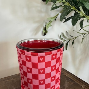 May include: A glass jar with a red interior, encased in a fabric sleeve. The sleeve displays a red and pink chequered pattern, each square featuring a white heart. The jar is placed on a wooden surface.