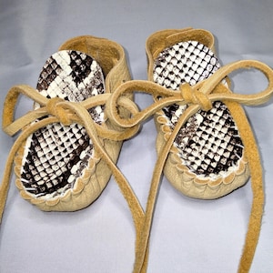Puede incluir: Un par de mocasines de bebé de cuero marrón con forro de estampado de serpiente blanco y negro y cordones de cuero marrón.