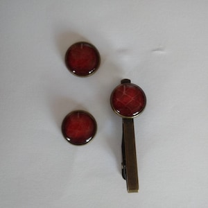 Puede incluir: Un conjunto de tres accesorios: dos gemelos redondos y un clip para corbata. Cada pieza presenta un diseño rojo oscuro con un marco de metal color bronce. El clip para corbata es rectangular.