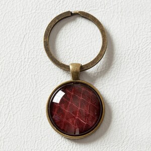 May include: A round keychain with a bronze-coloured metal ring and pendant. The pendant features a burgundy-coloured, textured design under a glass dome. The keychain is set against a white background.