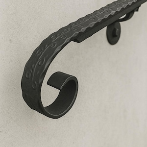 Puede incluir: Una barandilla de hierro forjado negro con una superficie texturizada y adornos decorativos. La barandilla está montada en una pared de color claro. El diseño presenta un extremo curvado y patrones decorativos en la parte superior.