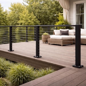 Könnte beinhalten: Eine moderne Außenterrasse mit einem schwarzen Metallgeländer und Kabelsystem. Die Terrasse hat eine braune Holzoberfläche und ist mit einem Korbmöbelsofa und Kissen ausgestattet. Grüne Pflanzen und Bäume sind im Hintergrund zu sehen.