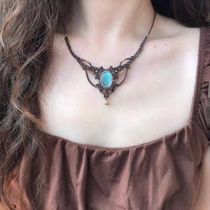 May include: A macrame necklace featuring a central oval blue gemstone set in dark brown cord. The design includes intricate knotted patterns, small gold-coloured beads integrated into the cord, and a single small gold-coloured drop pendant at the base.