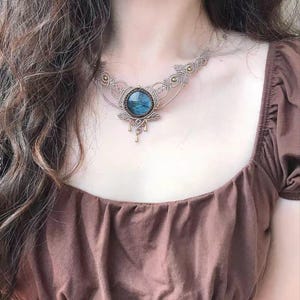 May include: A macrame necklace featuring a central round labradorite gemstone with blue iridescence, surrounded by intricate beige cord knotting, small metallic beads, and three small dangling metallic charms at the bottom of the pendant.