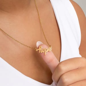 Könnte beinhalten: Eine goldfarbene Halskette mit dem Wort "hope" in kursiver Schrift. Die Halskette wird von einem Finger gehalten. Die Person trägt ein weißes Tanktop.