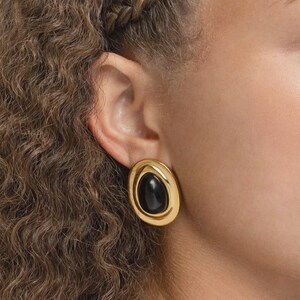 May include: Gold-toned oval earrings with a black stone center. The earrings have a polished finish and a classic design. The black stone is smooth and contrasts with the gold frame. These earrings are a stylish accessory.