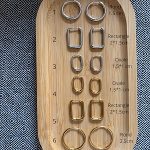Peut inclure: Une collection de six paires de boucles d'oreilles créoles disposées sur un plateau en bois. L'ensemble comprend des boucles argentées et dorées de formes ronde, rectangulaire et ovale, avec leurs dimensions indiquées en centimètres.