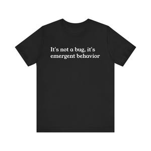 May include: Black t-shirt with the white text: "It's not a bug, it's emergent behavior." The shirt has a crew neck and short sleeves. The text is centered on the front of the shirt.