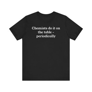 May include: Black t-shirt with the white text "Chemists do it on the table - periodically." The shirt has short sleeves and a crew neck. The text is centered on the front of the shirt.
