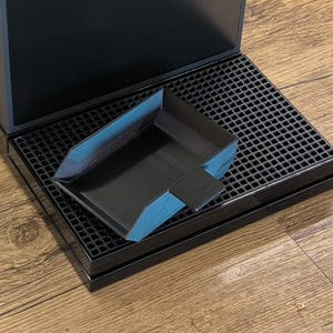 May include: A black, angled tray with blue accents rests on a black, grid-patterned surface. The tray is positioned on top of a black rectangular base, with a larger black structure in the background. The items are on a wooden surface.