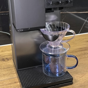Könnte beinhalten: Eine schwarze Kaffeemaschine mit digitaler Anzeige "60 16". Ein klarer, kegelförmiger Filter befindet sich über einer transparenten Glas-Tasse mit blauem Griff. Die Maschine steht auf einem schwarzen Sockel.