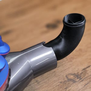 May include: Close-up of a vacuum cleaner attachment. The attachment is composed of a silver-gray body and a black, curved nozzle. The blue and red motor housing is visible in the background. The attachment is designed to connect to the vacuum cleaner.
