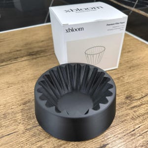 Può includere: Un portafiltro per caffè nero con un design scanalato è posto davanti a una scatola bianca. La scatola è etichettata "xbloom" e "Premium Filter Paper". Il supporto è progettato per contenere filtri di carta per preparare il caffè. La scatola contiene 100 fogli di filtri di carta.