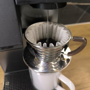May include: A close-up of a coffee brewing setup. Water streams from a black machine into a stainless steel pour-over filter with a paper filter. The filter sits atop a white mug with a brown handle, ready to receive the freshly brewed coffee.