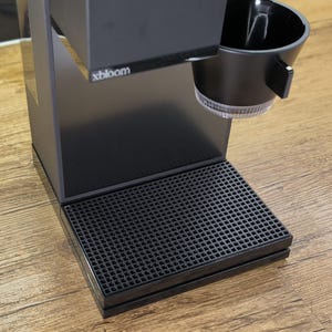 May include: A black coffee maker with the brand name "xbloom" on the front. The machine has a black drip tray and a black cup holder. The coffee maker is on a wooden surface.