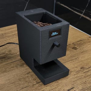 Könnte beinhalten: Ein schwarzer Kaffeebohnenzähler mit digitaler Anzeige "Bean Counter". Das Gerät hat einen Knopf und eine kleine Ablage unten. Der obere Bereich enthält Kaffeebohnen.