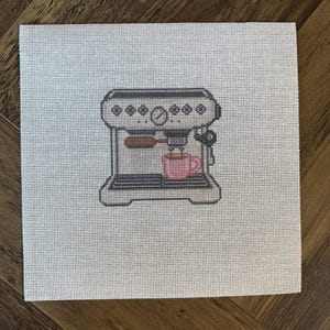 May include: A square needlepoint canvas featuring a centered, detailed design of a grey espresso machine with a brown portafilter handle and a small pink coffee cup placed on the drip tray, all rendered in a grid-like stitch pattern.