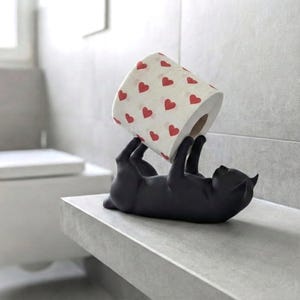 Könnte beinhalten: Ein mattschwarzer Toilettenpapierhalter in Katzenform, der auf dem Rücken liegt und mit den Pfoten eine Rolle weißes Toilettenpapier mit einem wiederkehrenden Muster aus kleinen roten Herzen hält. Die Katzenfigur ist schwarz und hat spitze Ohren.