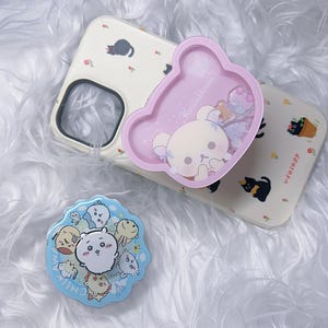 May include: A beige phone case with a black cat and flower pattern, a pink bear-shaped phone grip with "Korilakkuma" text, and a blue circular phone grip with cartoon characters.