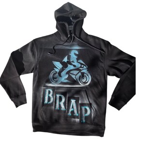 Pode incluir: Moletom preto com capuz com um gráfico de motocicleta azul claro e a palavra "BRAP". O design apresenta um piloto em silhueta em uma moto de corrida. O moletom tem capuz com cordão e um bolso frontal.
