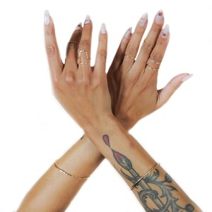 Pu&ograve; includere: Due mani incrociate all'altezza dei polsi, con anelli d'oro alle dita. La mano sinistra ha un anello quadrato sul dito medio. La mano destra ha diversi anelli sottili d'oro alle dita. Entrambe le mani hanno smalto per unghie bianco con glitter. La mano destra ha un tatuaggio di un pennello rosso sul polso.