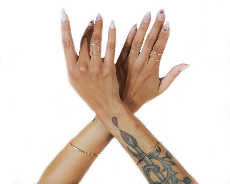Pu&ograve; includere: Due mani incrociate all'altezza dei polsi, con anelli d'oro alle dita. La mano sinistra ha un anello quadrato sul dito medio. La mano destra ha diversi anelli sottili d'oro alle dita. Entrambe le mani hanno smalto per unghie bianco con glitter. La mano destra ha un tatuaggio di un pennello rosso sul polso.