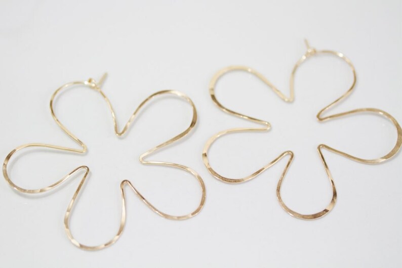 May include: A pair of gold flower-shaped hoop earrings. The earrings are made of thin, hammered metal and have a delicate, feminine look.