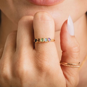 Könnte beinhalten: Ein goldener Ring mit fünf farbigen Edelsteinen. Der Ring wird an einem Finger getragen, wodurch die Farben der Edelsteine hervorgehoben werden. Ein weiterer goldener Ring und eine goldene Halskette sind ebenfalls zu sehen.