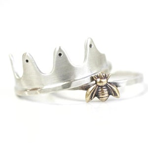 May include: Two silver rings, one with a crown design and the other with a small bee charm. The bee charm is made of gold and has a textured surface.