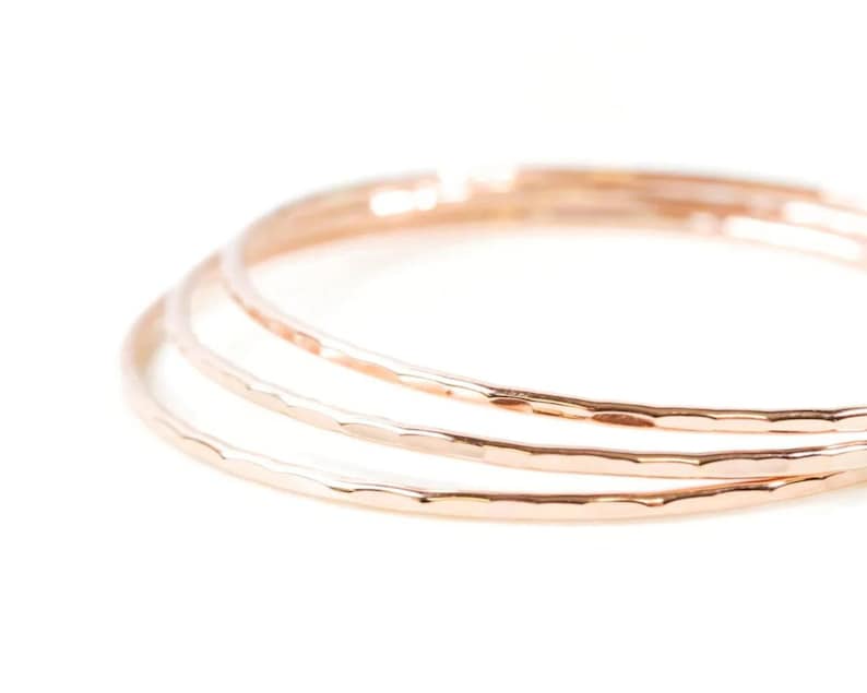 May include: Three thin, hammered gold rings stacked together. The rings are smooth and shiny, and they have a delicate, feminine look.
